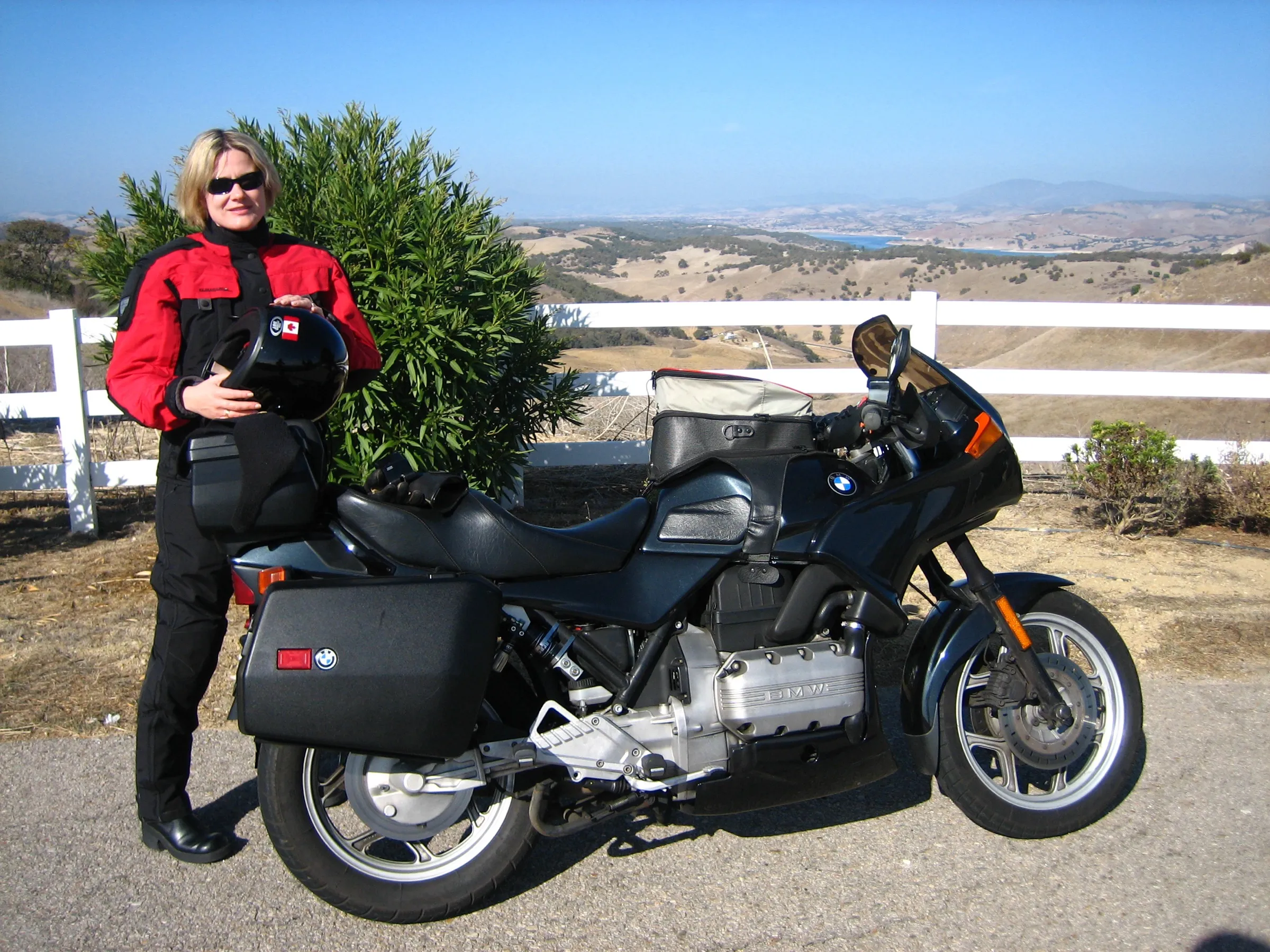 Sharon in red and black riding gear holding her helmet beside the BMW K75S
