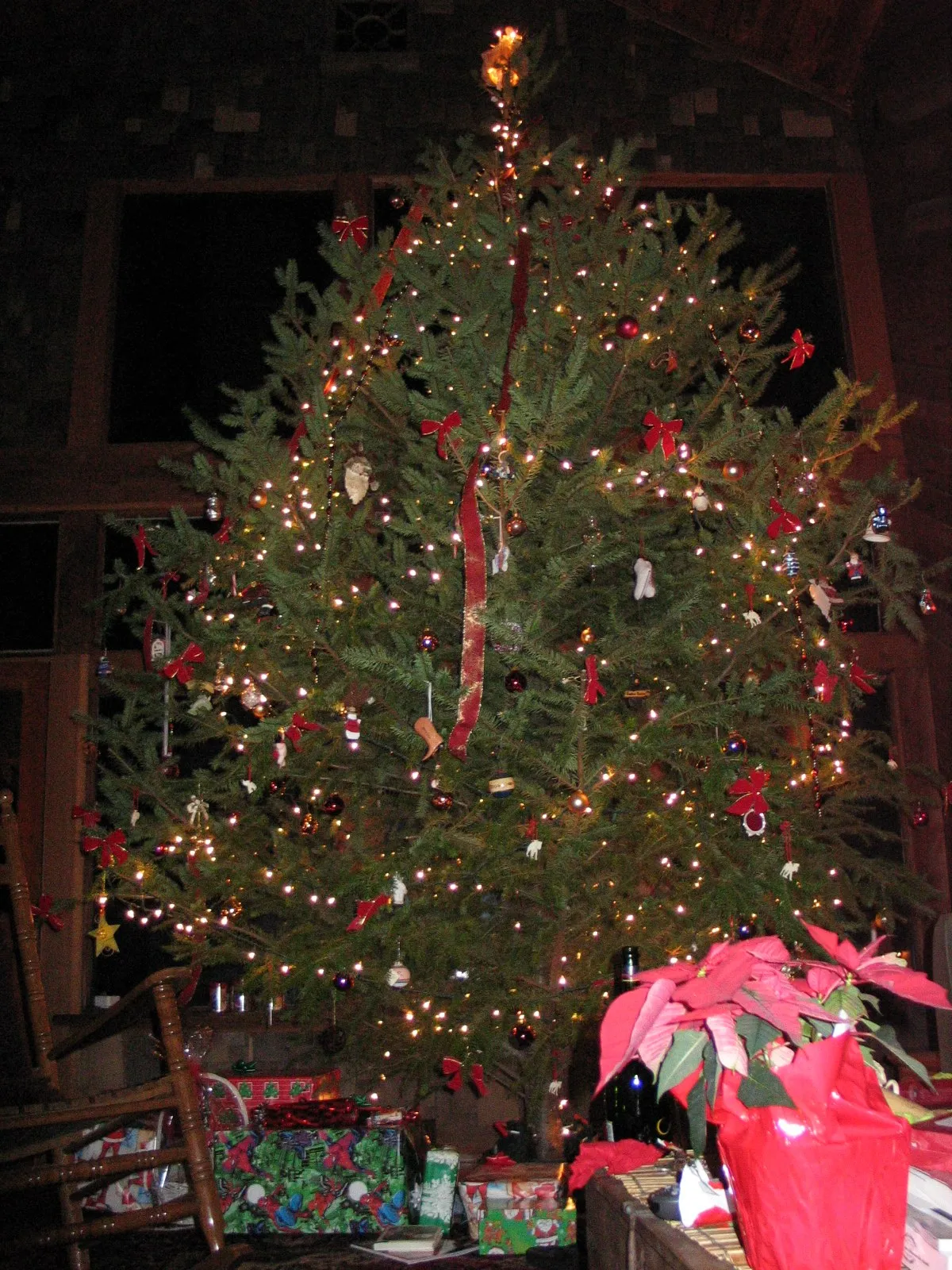 Tall Christmas tree with lights and red bows beside a pile of wrapped gifts